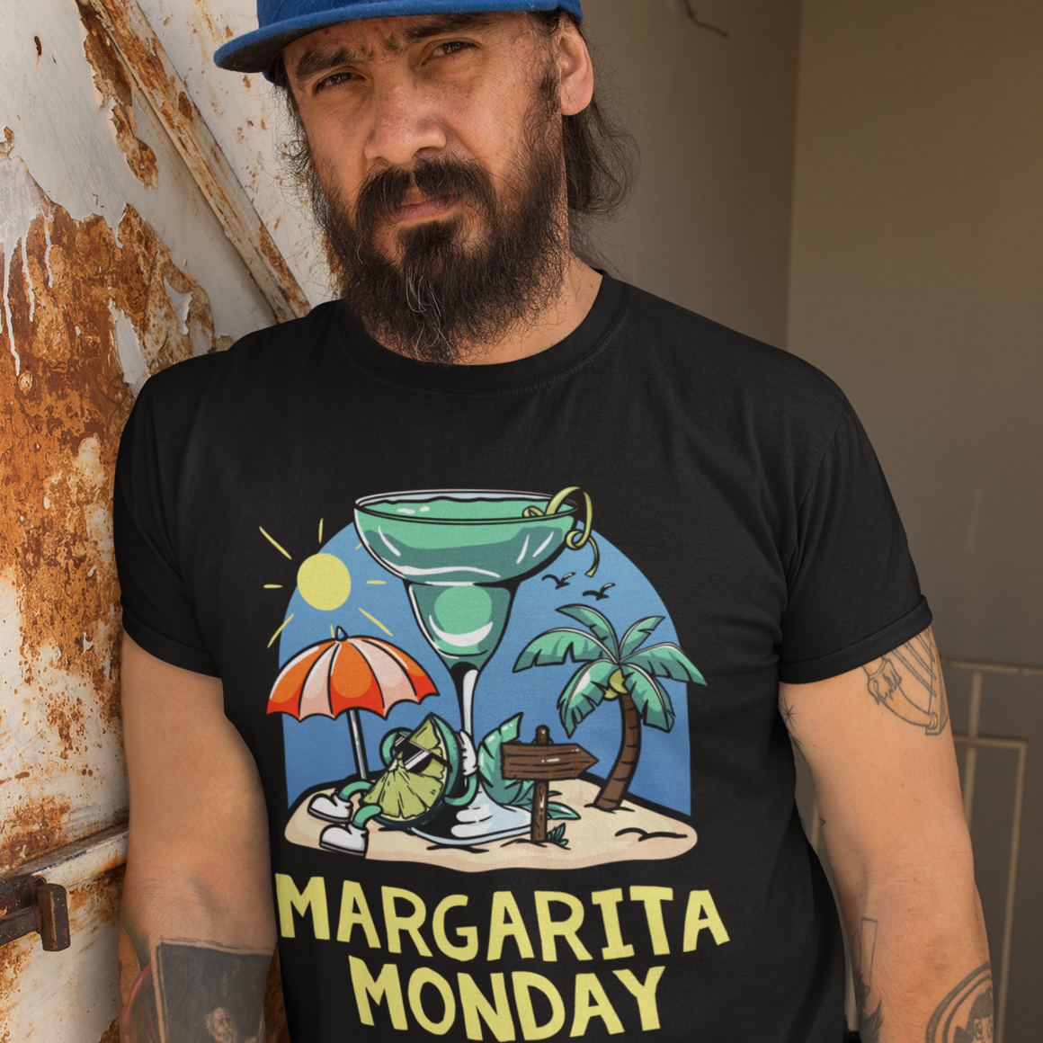 Margarita Monday T-shirt Design Ideas featuring a black t-shirt with a colorful tropical design of a margarita glass, palm trees, a sun umbrella, and a sandy beach scene, complemented by bold yellow text reading 'Margarita Monday.' Modeled by a bearded man in a blue cap, adding a casual and relaxed vibe.