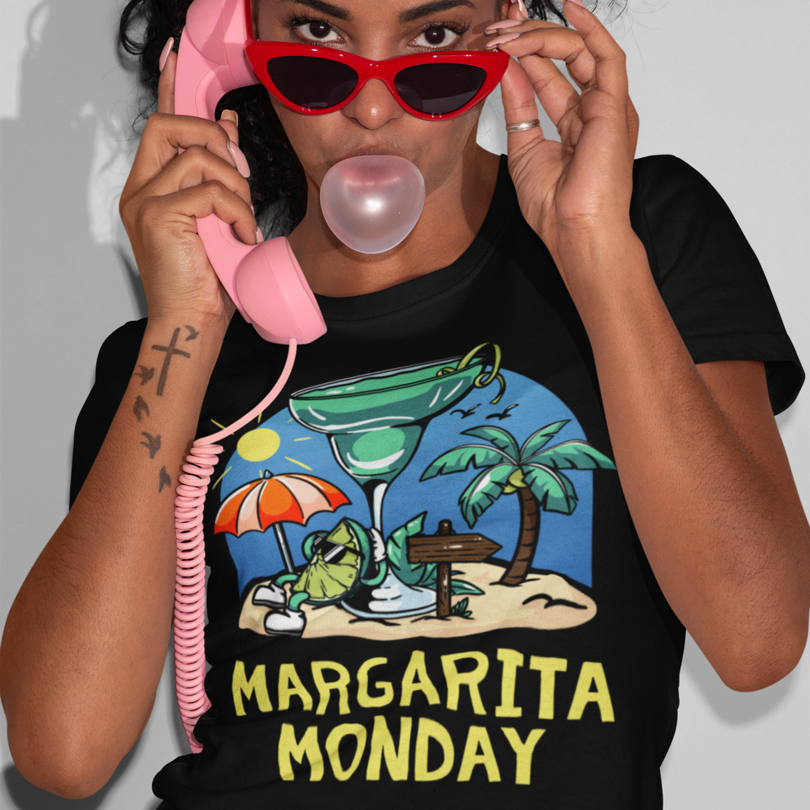 Margarita Monday T-shirt Design Ideas featuring a black t-shirt with a vibrant tropical design of a margarita glass, palm trees, a sun umbrella, and a sandy beach scene, highlighted by bold yellow text reading 'Margarita Monday.' Modeled by a stylish woman with red sunglasses and a playful pose.