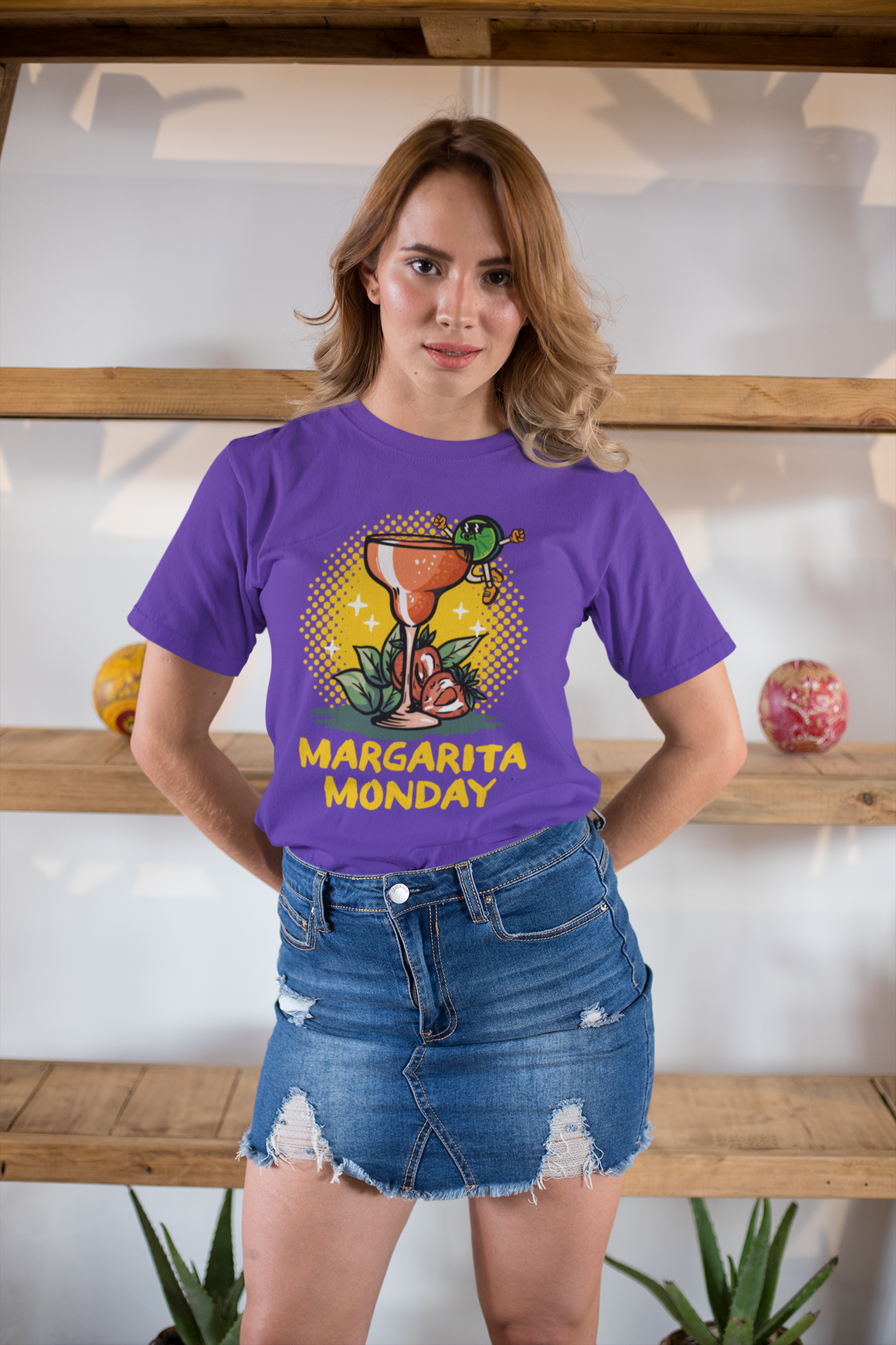 Woman wearing a purple T-shirt with a "Margarita Monday" design, featuring a vibrant margarita glass illustration and bold yellow text. This trendy Margarita Monday T-shirt style adds a fun and festive touch to her casual outfit, perfect for celebrating Margarita Monday with style. The background shelves add a cozy, rustic vibe.