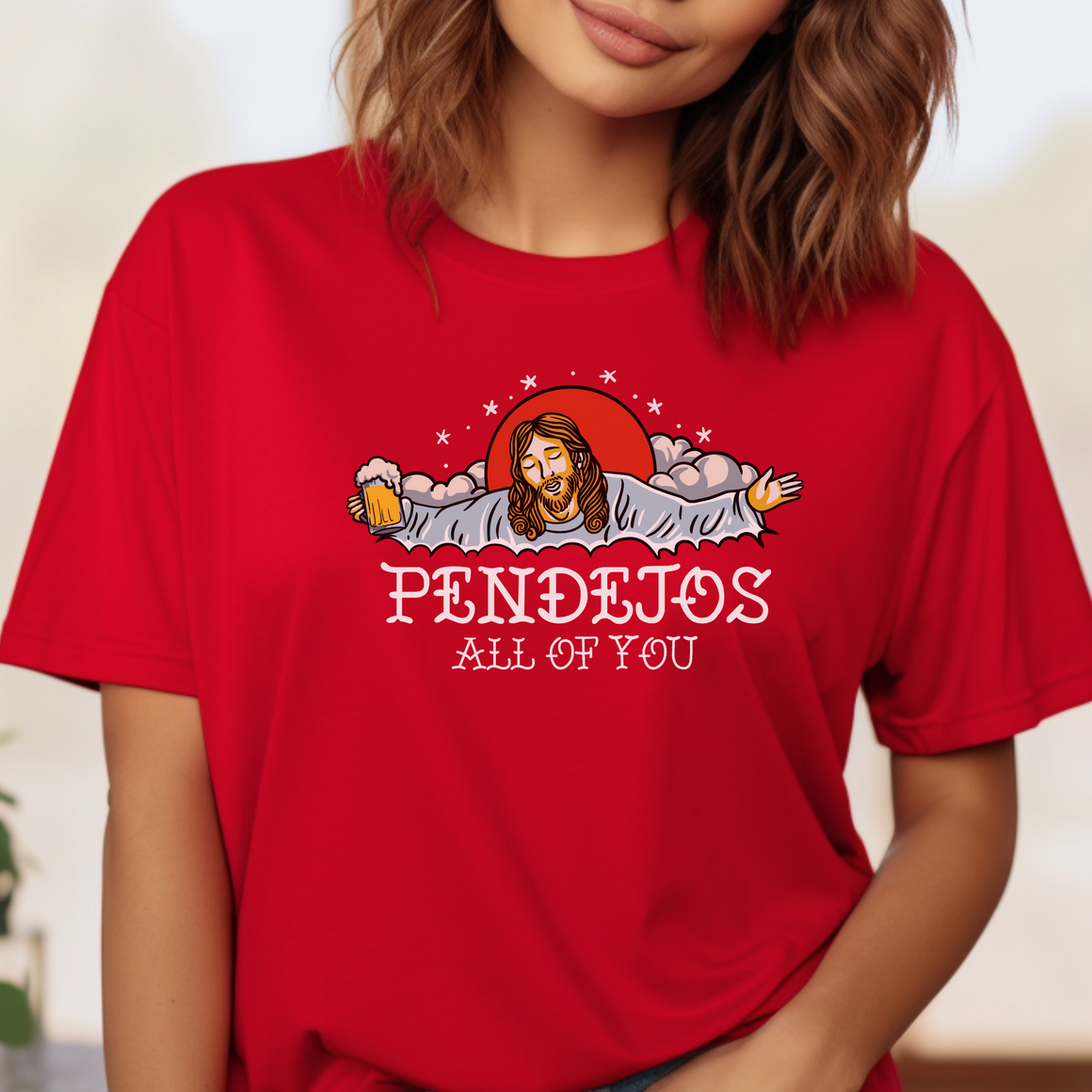 A person wearing a red t-shirt featuring an illustration of a bearded figure with open arms, holding a beer mug, surrounded by clouds and a red sun. The text "PENDEJOS ALL OF YOU" is printed below the image.