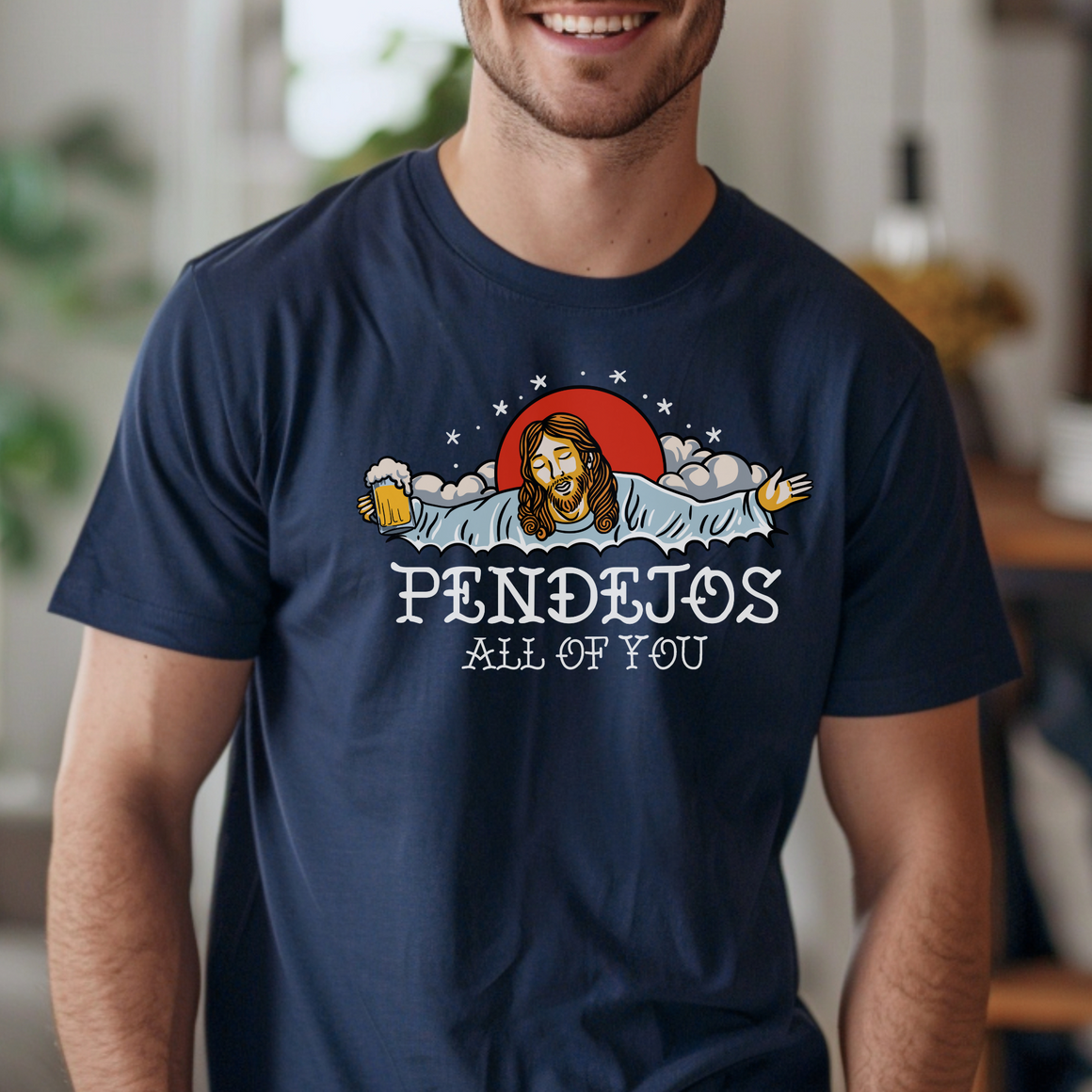 A man wearing a navy blue t-shirt featuring an illustration of a bearded figure with open arms, holding a beer mug, surrounded by clouds and a red sun. The text "PENDEJOS ALL OF YOU" is printed below the image.
