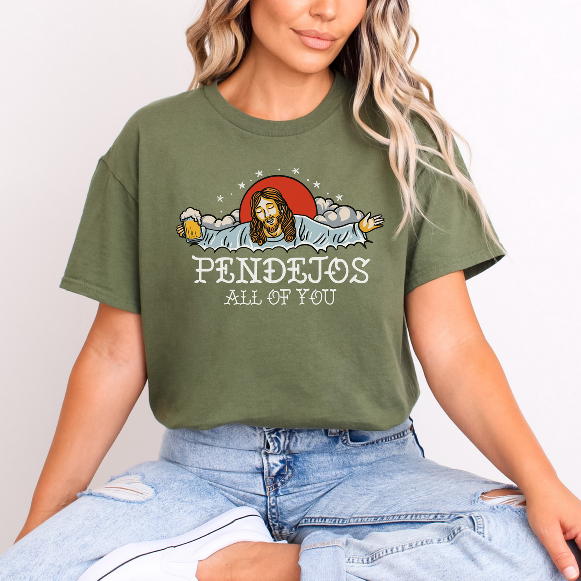 Person wearing an olive green t-shirt with a humorous graphic of a figure resembling Jesus holding a beer mug, surrounded by clouds and stars. The text on the shirt reads 'Pendejos All of You.' The person is seated cross-legged, wearing light blue jeans."