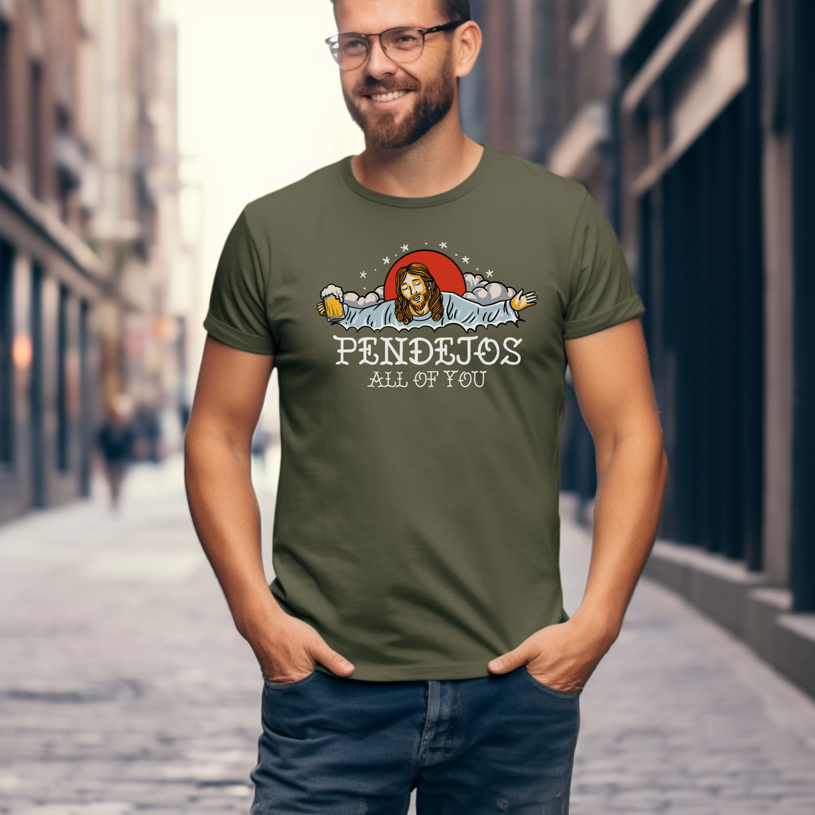 Man standing on a city street wearing an olive green t-shirt with a humorous graphic of a figure resembling Jesus holding a beer mug, surrounded by clouds and stars. The text on the shirt reads 'Pendejos All of You.' The man is smiling, with his hands in his pockets.