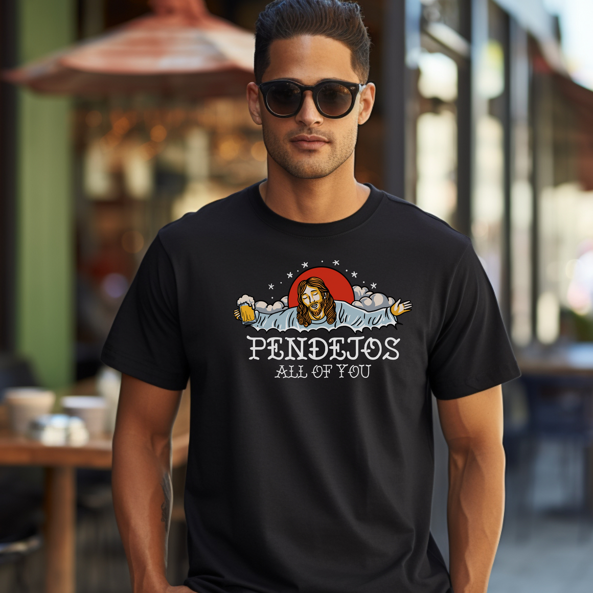 Man wearing sunglasses and a black t-shirt with a humorous graphic of a figure resembling Jesus holding a beer mug, surrounded by clouds and stars. The text on the shirt reads 'Pendejos All of You.' The man is standing outdoors in a casual setting.