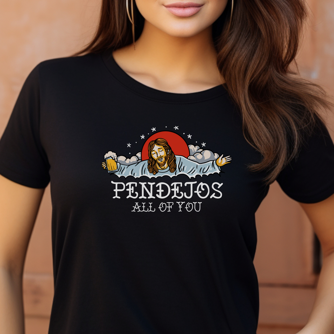 Person wearing a black t-shirt featuring a humorous graphic of a figure resembling Jesus holding a beer mug, surrounded by clouds and stars. The text on the shirt reads 'Pendejos All of You.' The person is standing against a warm-colored background.