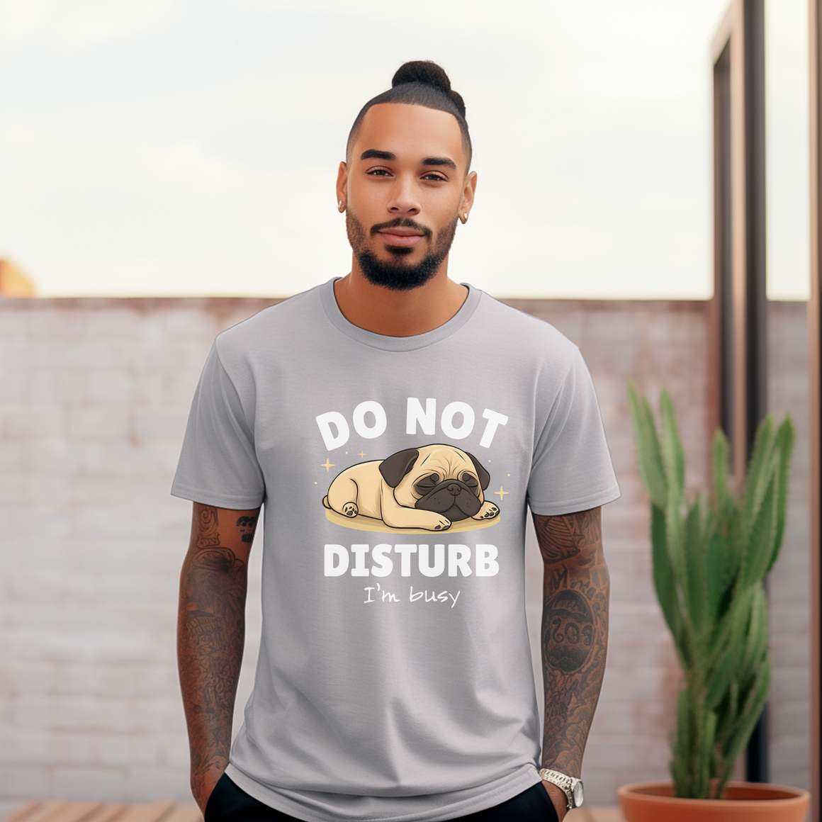 A man wearing a gray t-shirt with a cute sleeping pug illustration and the text "DO NOT DISTURB, I'm busy" printed on it. He has tattoos on his arms and stands outdoors near a cactus.