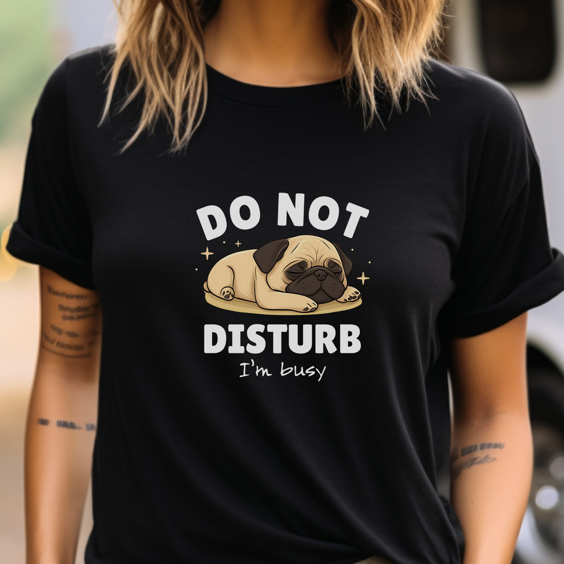 A person wearing a black t-shirt with a cute sleeping pug illustration and the text "DO NOT DISTURB, I'm busy" printed on it. The person has shoulder-length hair and visible tattoos on their arm.