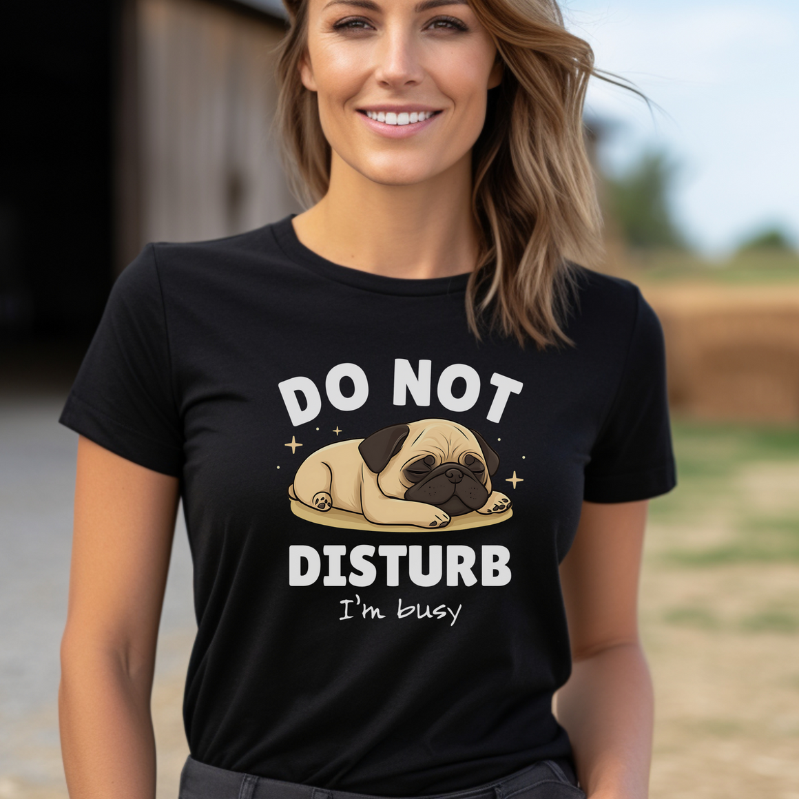 A woman wearing a black t-shirt with a cute sleeping pug illustration and the text "DO NOT DISTURB, I'm busy" printed on it. She is smiling and standing outdoors with a barn and field in the background.