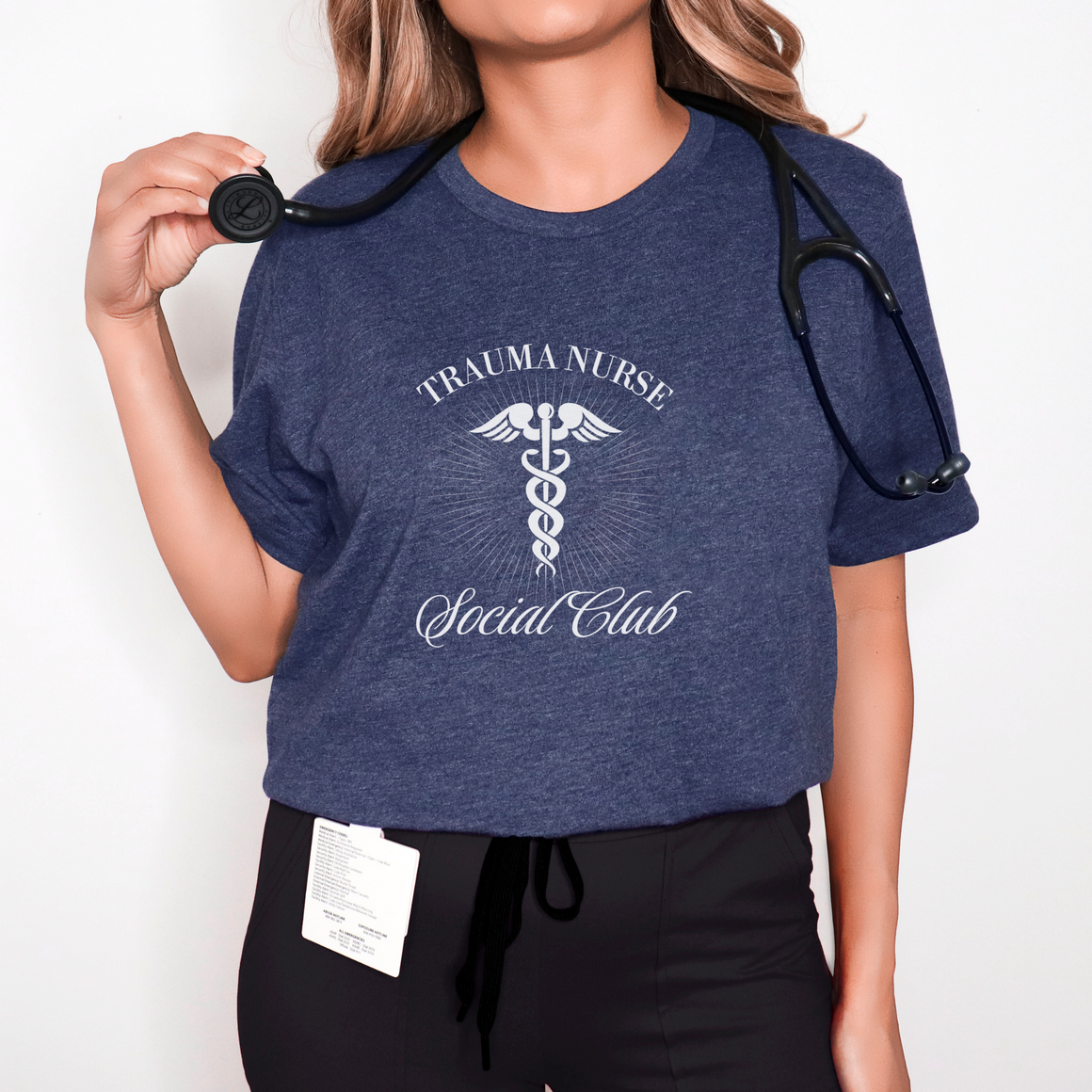 A nurse standing wearing a shirt that says Trauma Nurse Social Club and has a grapgic of a nurses caduceus