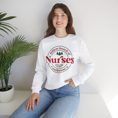 White Christmas sweatshirt with the humorous phrase 'Jolliest bunch of Nurses this side of the nuthouse,' surrounded by festive elements like trees and holly. A perfect holiday-themed gift for nurses with a sense of humor, celebrating Christmas in a fun and lighthearted style.