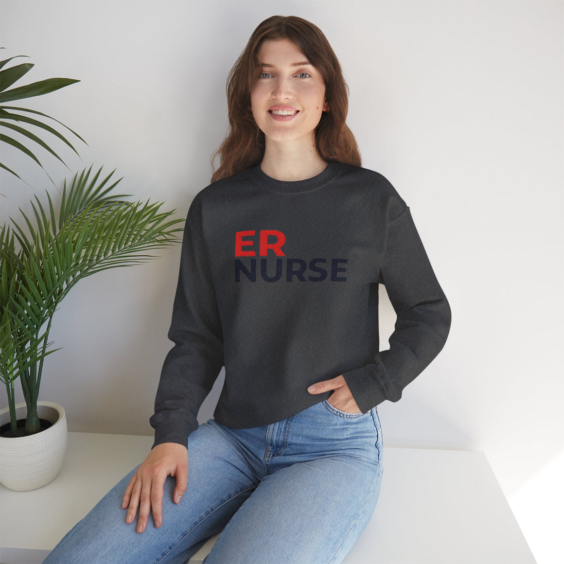 A woman smiling and wearing a charcoal ER Nurse Sweatshirt, sitting casually with a potted plant beside her.