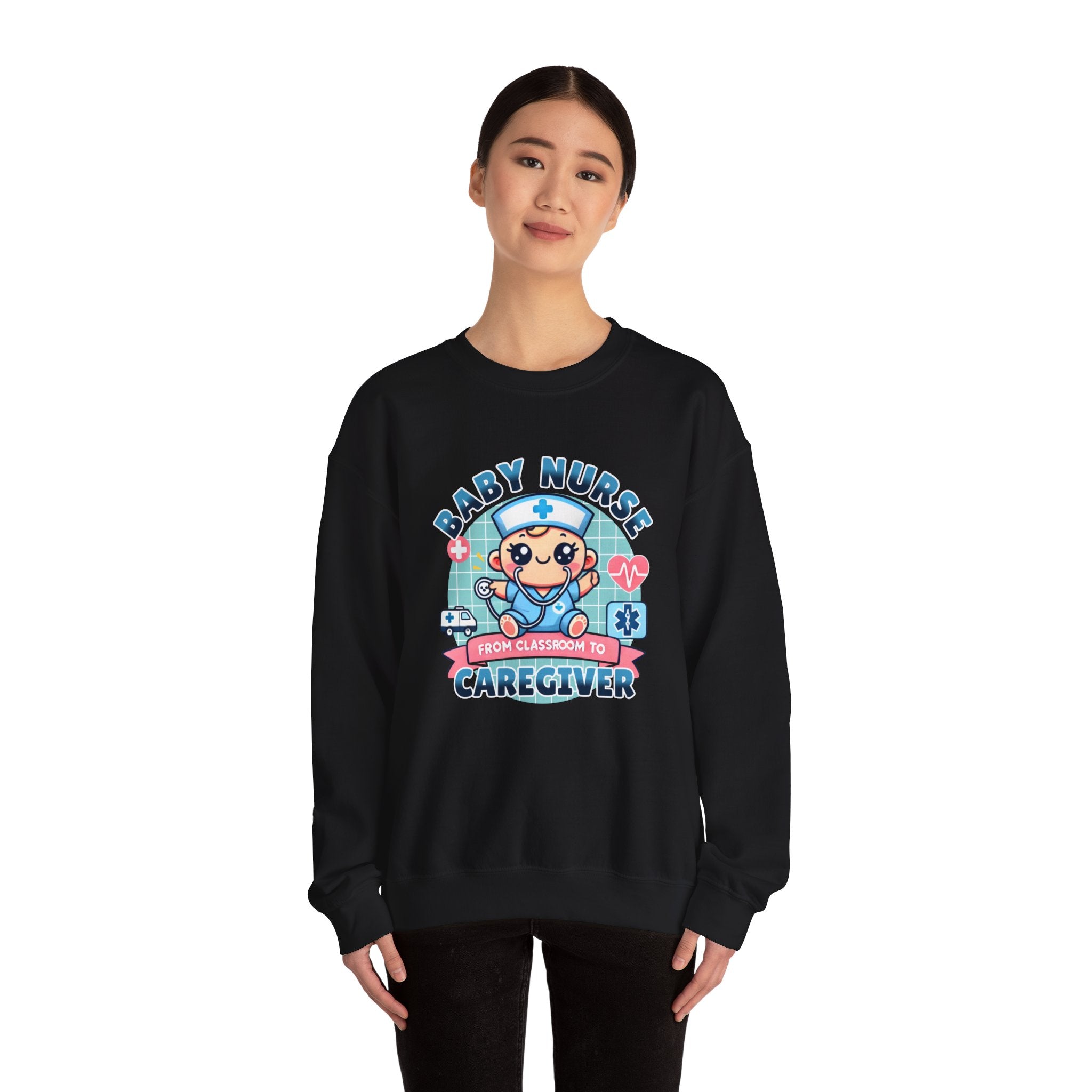 Woman wearing a black sweatshirt with a cute, cartoon-style design that reads 'Baby Nurse - From Classroom to Caregiver.' The graphic features a smiling baby nurse character in scrubs, holding a stethoscope, with icons of medical elements like an ambulance, heartbeat, and medical cross around. The woman holds a coffee mug in one hand and has a stethoscope around her neck.