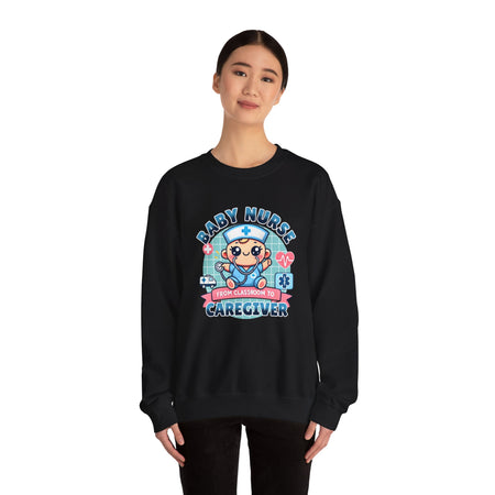 Woman wearing a black sweatshirt with a cute, cartoon-style design that reads 'Baby Nurse - From Classroom to Caregiver.' The graphic features a smiling baby nurse character in scrubs, holding a stethoscope, with icons of medical elements like an ambulance, heartbeat, and medical cross around. The woman holds a coffee mug in one hand and has a stethoscope around her neck.