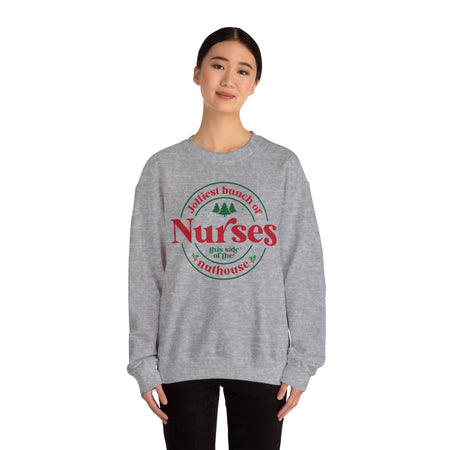 Grey Christmas sweatshirt with the humorous phrase 'Jolliest bunch of Nurses this side of the nuthouse,' surrounded by festive elements like trees and holly. A perfect holiday-themed gift for nurses with a sense of humor, celebrating Christmas in a fun and lighthearted style.