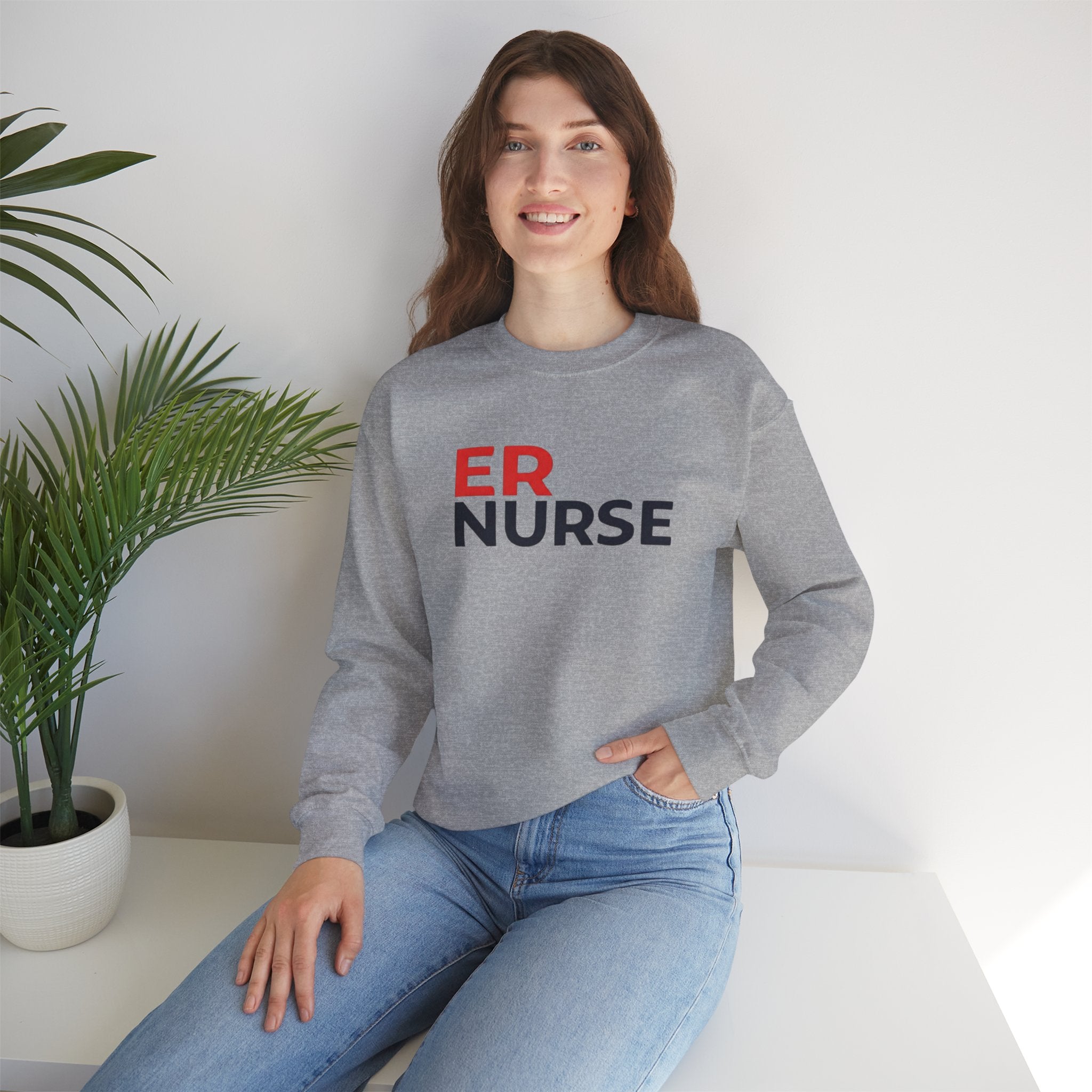 A woman smiling and wearing a light gray ER Nurse Sweatshirt, sitting casually with a potted plant beside her.