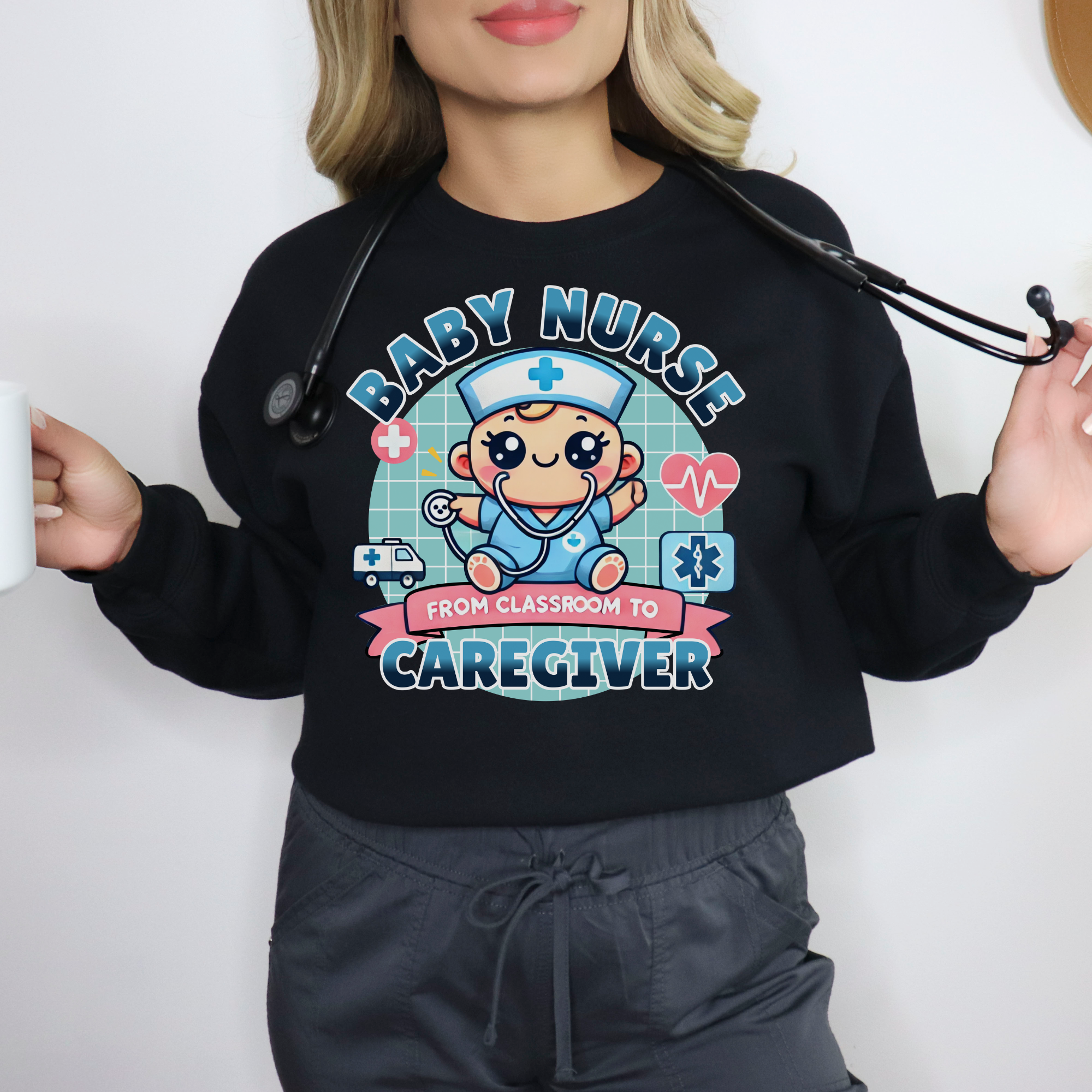 Woman wearing a black sweatshirt with a cute, cartoon-style design that reads 'Baby Nurse - From Classroom to Caregiver.' The graphic features a smiling baby nurse character in scrubs, holding a stethoscope, with icons of medical elements like an ambulance, heartbeat, and medical cross around. The woman holds a coffee mug in one hand and has a stethoscope around her neck.