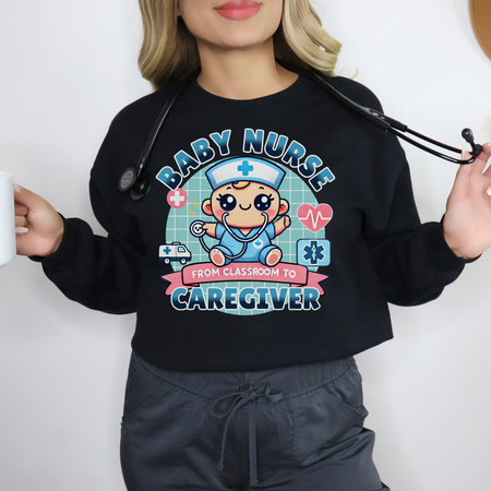 Woman wearing a black sweatshirt with a cute, cartoon-style design that reads 'Baby Nurse - From Classroom to Caregiver.' The graphic features a smiling baby nurse character in scrubs, holding a stethoscope, with icons of medical elements like an ambulance, heartbeat, and medical cross around. The woman holds a coffee mug in one hand and has a stethoscope around her neck.