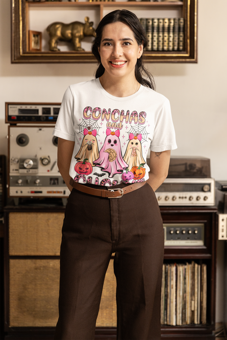 Woman wearing a white t-shirt featuring the "Conchas and Cucuy's" design, part of our Latino-inspired Halloween apparel. The shirt displays three cute ghost figures holding conchas and decorated with pink bows, spider webs, and festive pumpkin accents, combining Halloween spirit with Día de los Muertos charm. This unisex, soft-style shirt offers a unique blend of spooky and sweet for the Halloween season.