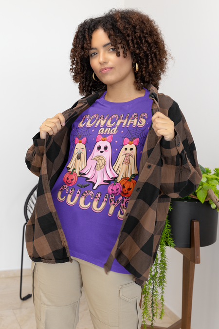 Woman wearing a purple t-shirt featuring the "Conchas and Cucuy's" design, part of our Latino-inspired Halloween apparel. The shirt displays three cute ghost figures holding conchas and decorated with pink bows, spider webs, and festive pumpkin accents, combining Halloween spirit with Día de los Muertos charm. This unisex, soft-style shirt offers a unique blend of spooky and sweet for the Halloween season.