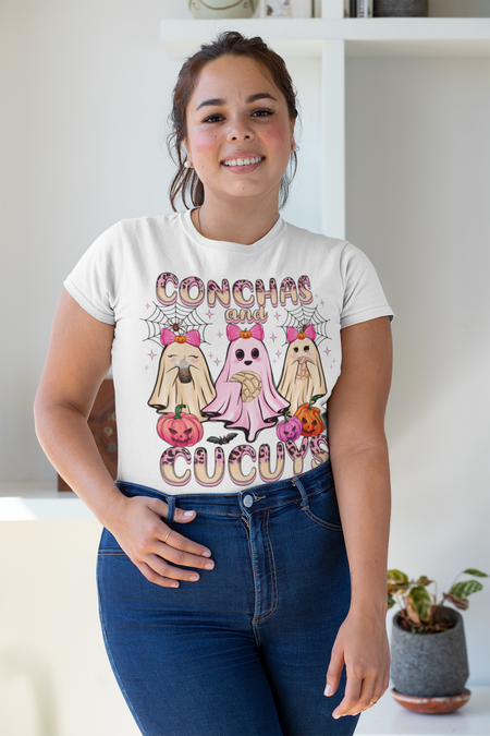 Woman wearing a white t-shirt featuring the "Conchas and Cucuy's" design, part of our Latino-inspired Halloween apparel. The shirt displays three cute ghost figures holding conchas and decorated with pink bows, spider webs, and festive pumpkin accents, combining Halloween spirit with Día de los Muertos charm. This unisex, soft-style shirt offers a unique blend of spooky and sweet for the Halloween season.