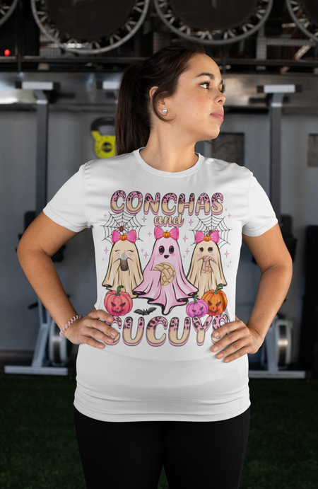Woman wearing a white t-shirt featuring the "Conchas and Cucuy's" design, part of our Latino-inspired Halloween apparel. The shirt displays three cute ghost figures holding conchas and decorated with pink bows, spider webs, and festive pumpkin accents, combining Halloween spirit with Día de los Muertos charm. This unisex, soft-style shirt offers a unique blend of spooky and sweet for the Halloween season.