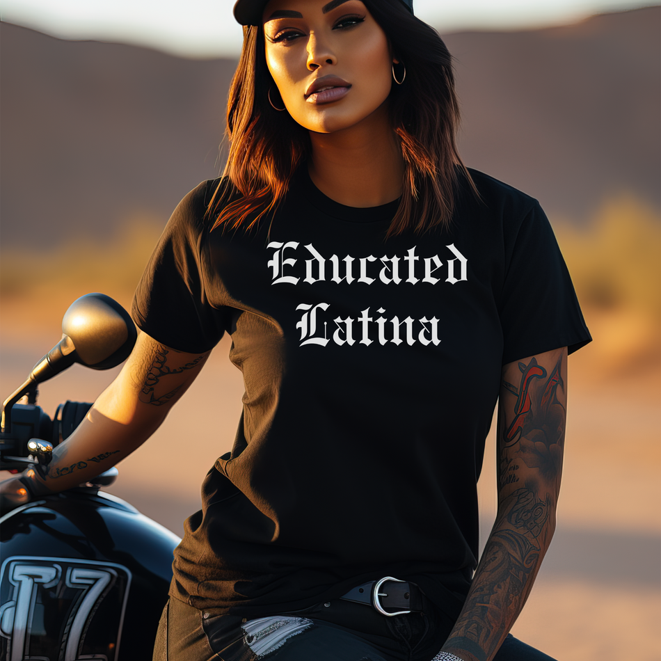 Confident woman with tattoos leaning on a motorcycle in the desert, wearing a black t-shirt that reads 'Educated Latina' in bold old English font. This shirt highlights educated Latina T-shirt sayings and quotes, celebrating Latina pride and empowerment in a stylish, edgy way. Perfect for showcasing cultural identity with confidence and flair.