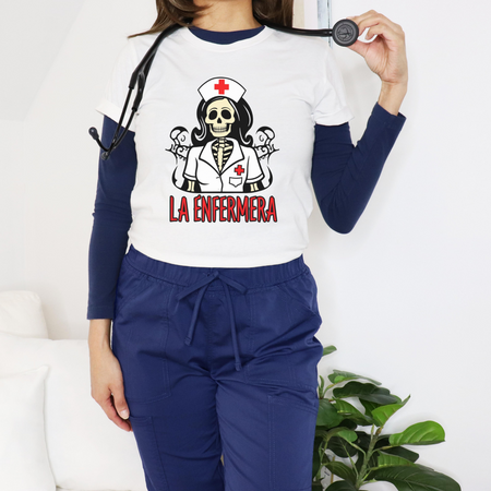 Woman dressed in medical scrubs, wearing a "La Enfermera Loteria Shirt" featuring a skeleton nurse in a white uniform with a red cross on the cap, holding a stethoscope. The design resembles a loteria card with "La Enfermera" in bold red letters, capturing a Día de los Muertos and healthcare theme.