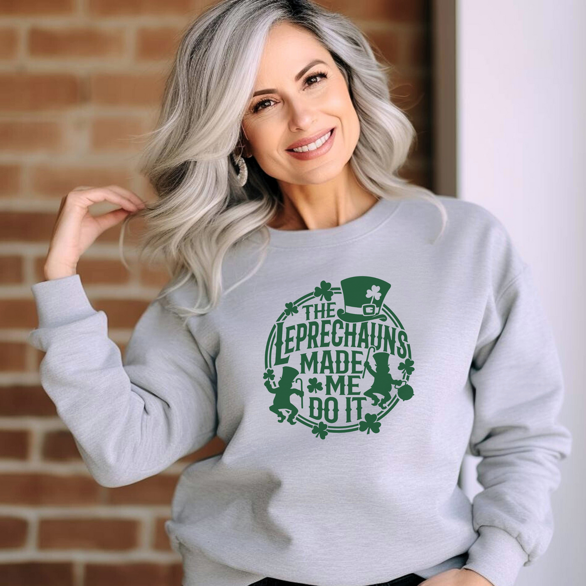 A woman with gray hair smiling and wearing a light gray St. Patrick's Day sweatshirt with a green design featuring leprechauns and the text 'THE LEPRECHAUNS MADE ME DO IT,' standing against a brick wall