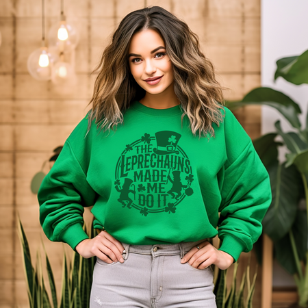A woman with wavy hair wearing a green St. Patrick's Day sweatshirt with a design featuring leprechauns and the text 'THE LEPRECHAUNS MADE ME DO IT,' standing in a plant-filled room.