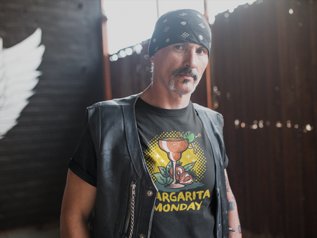 Man wearing a black T-shirt with a "Margarita Monday" design, featuring a colorful margarita glass graphic, layered under a leather vest and paired with a bandana. This trendy Margarita Monday T-shirt style adds a fun, festive twist to his edgy, biker-inspired look, perfect for Margarita Monday celebrations or casual outings. The rustic background enhances the rugged vibe.