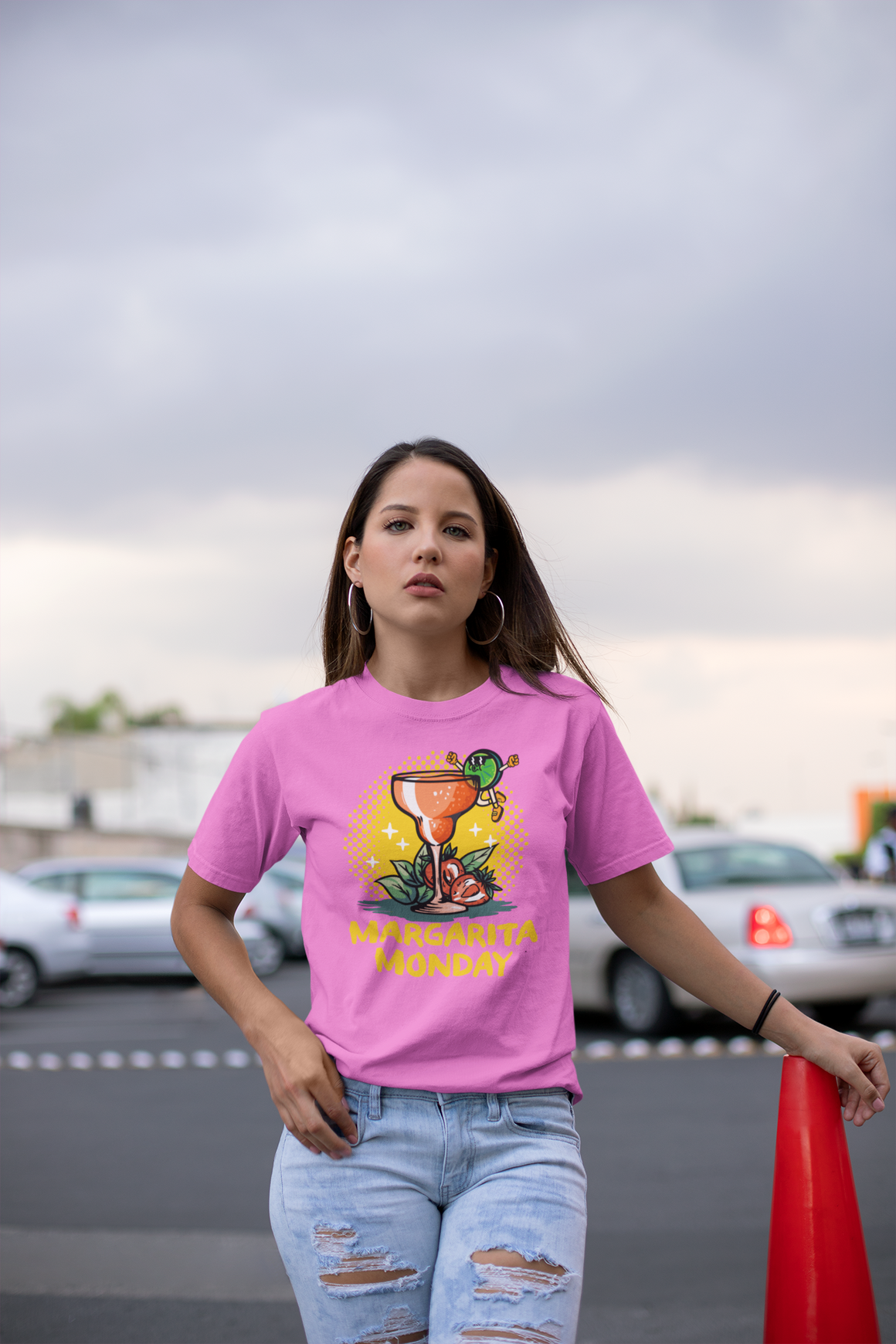 Woman wearing a pink T-shirt with a "Margarita Monday" design, featuring a colorful margarita glass graphic and bold lettering. This stylish Margarita Monday T-shirt design adds a fun and festive element to her casual look, perfect for celebrating Margarita Monday. The urban background enhances the relaxed, trendy vibe.