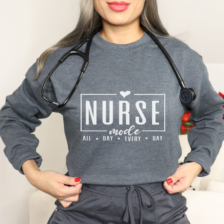 Woman wearing a gray sweatshirt with 'Nurse Mode All Day Every Day' printed in white text, a stethoscope around her neck, and hands on her waistband. Stylish design perfect for Nurses Week Custom Shirt Suggestions.