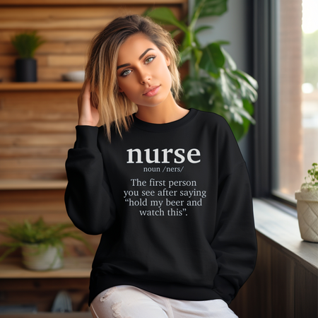 A woman wearing a black sweatshirt with a funny nurse-themed shirt design, reading: 'The first person you see after saying “hold my beer and watch this”.' She is sitting in a cozy room with plants in the background.