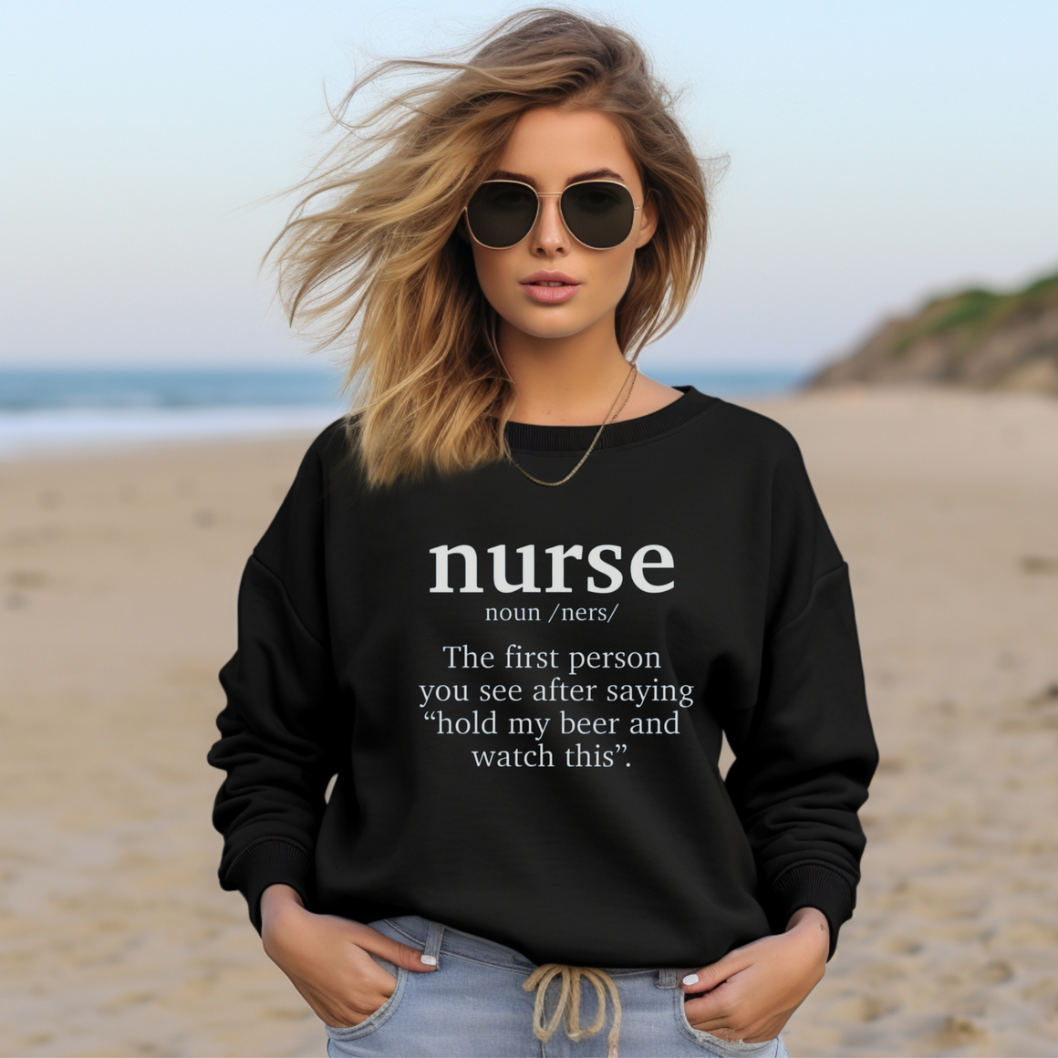 A woman wearing a black sweatshirt with a funny nurse-themed shirt design, reading: 'The first person you see after saying “hold my beer and watch this”.' She is sitting in a cozy room with plants in the background.