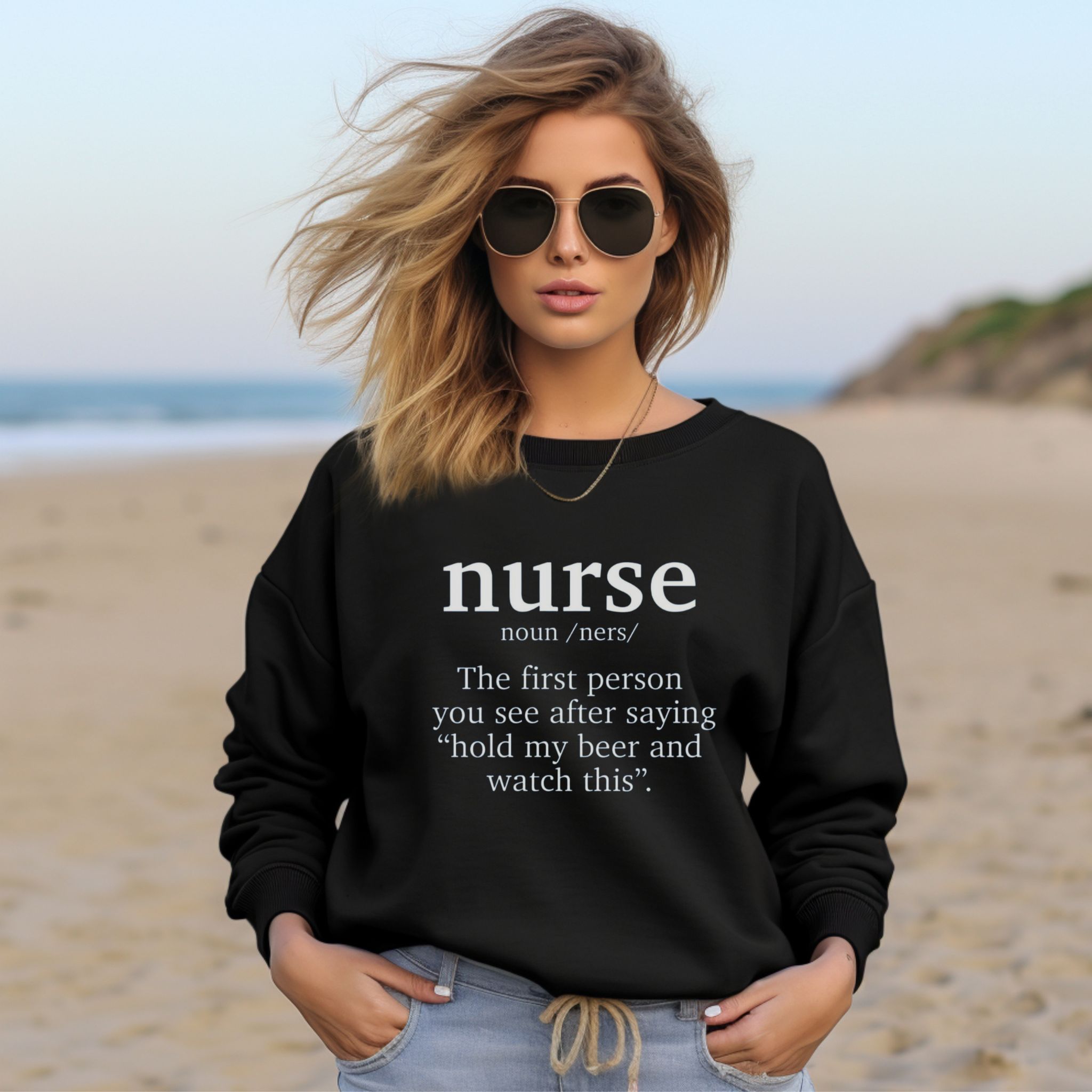 A woman wearing a black sweatshirt with a funny nurse-themed shirt design, reading: 'The first person you see after saying “hold my beer and watch this”.' She is sitting in a cozy room with plants in the background.