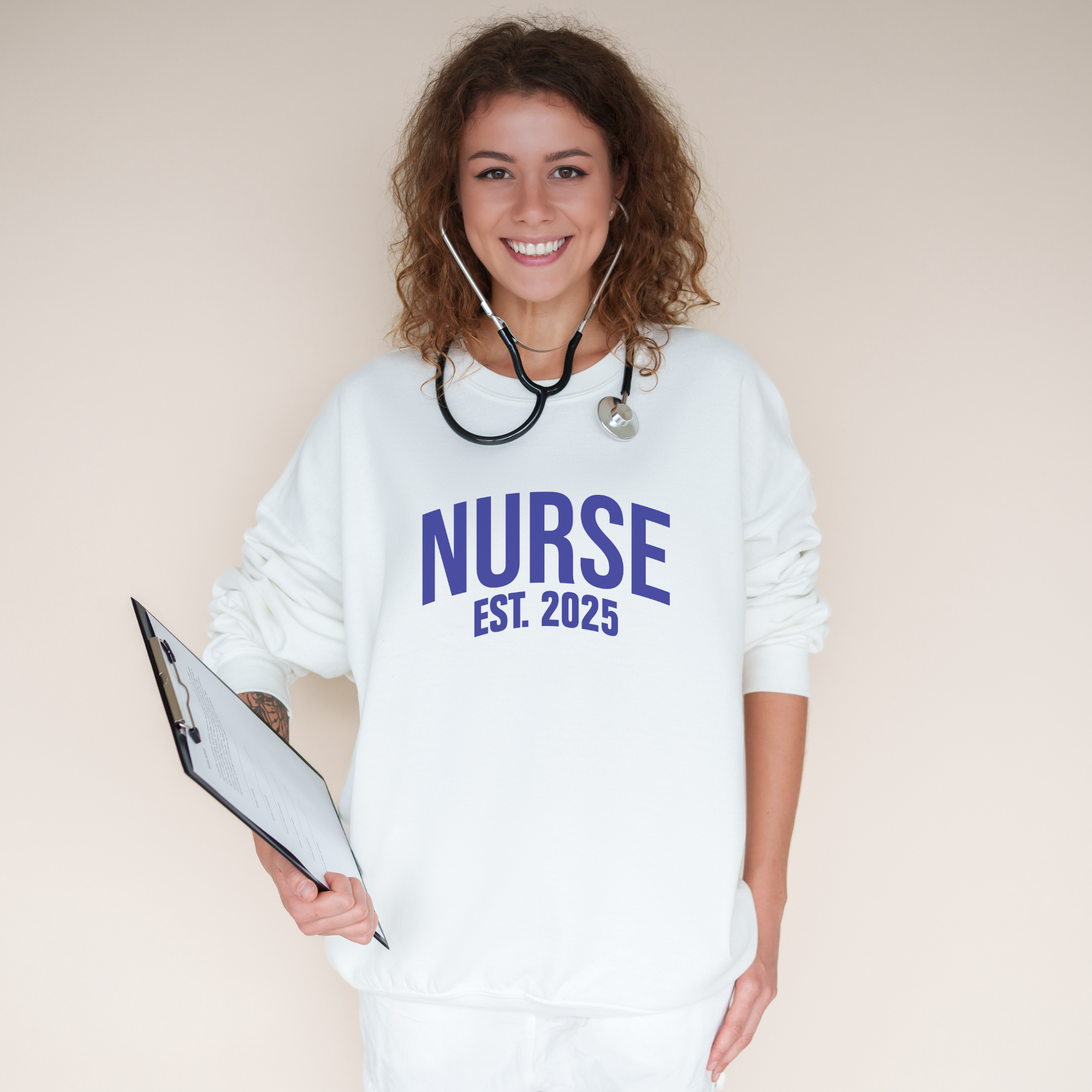 A smiling nurse wearing a "Nurse Est. 2025 Sweatshirt" in white with blue text, a stethoscope around her neck, and holding a clipboard.