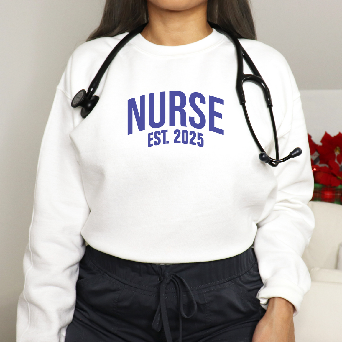 A nurse wearing a "Nurse Est. 2025 Sweatshirt" in white with blue text, a stethoscope around her neck, and dark drawstring pants.