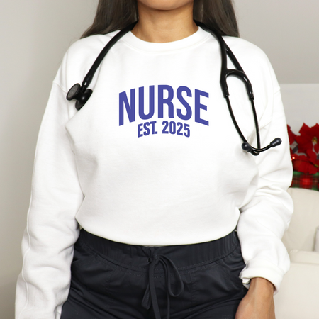 A nurse wearing a "Nurse Est. 2025 Sweatshirt" in white with blue text, a stethoscope around her neck, and dark drawstring pants.