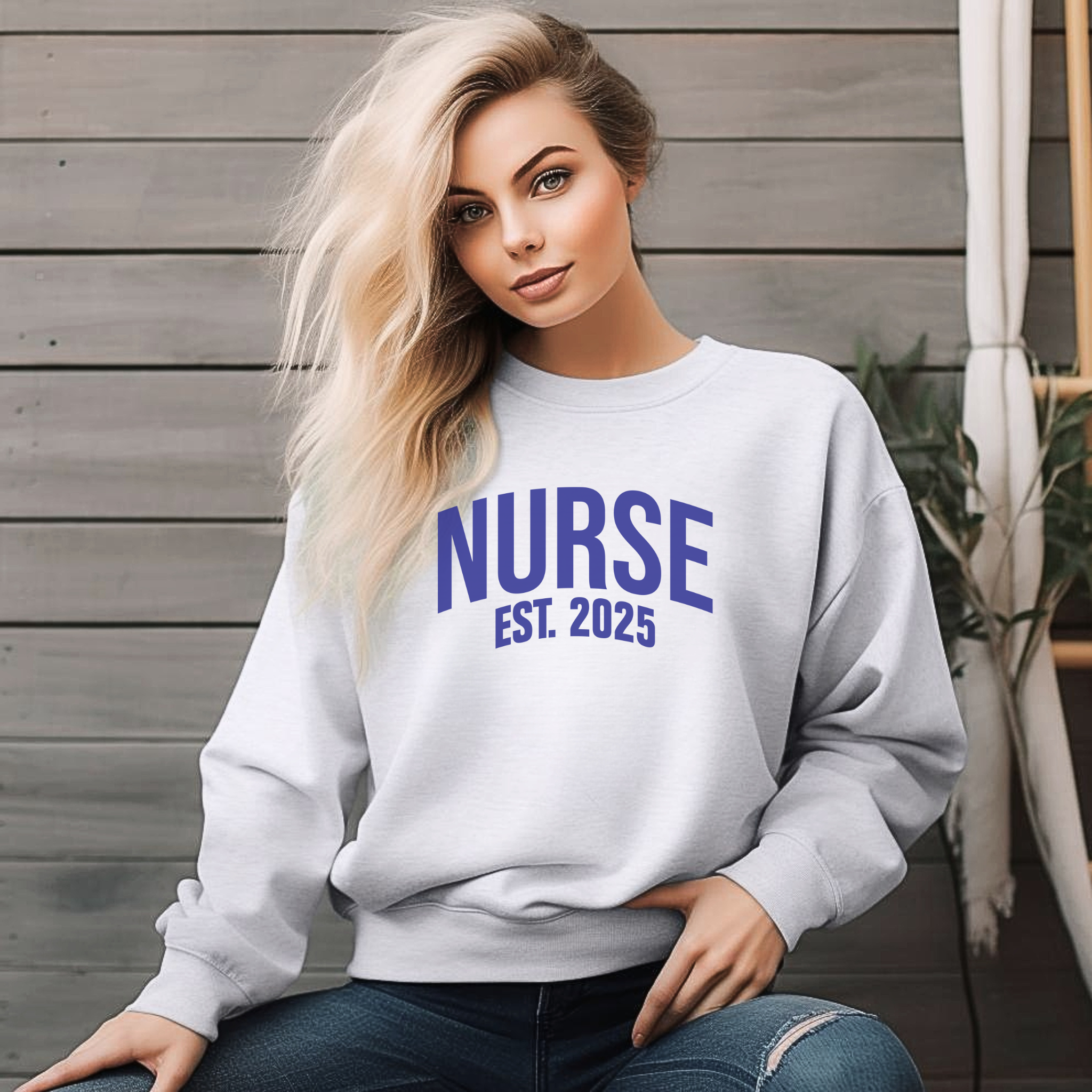 A woman with long blonde hair wearing a "Nurse Est. 2025 Sweatshirt" in light gray with blue text, seated outdoors against a wooden wall, looking relaxed.