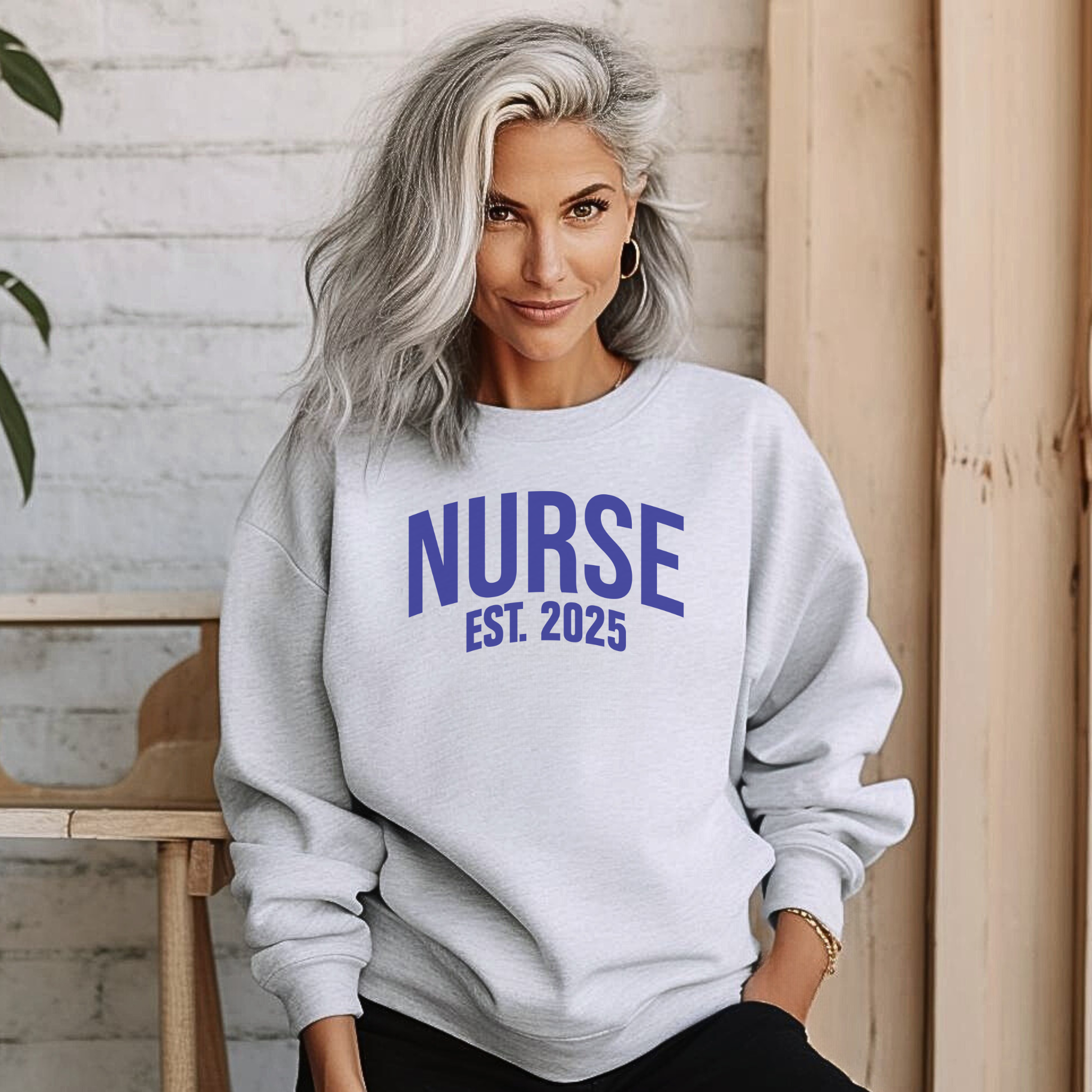 A nurse wearing a "Nurse Est. 2025 Sweatshirt" in white with blue text, a stethoscope around her neck, and dark drawstring pants.