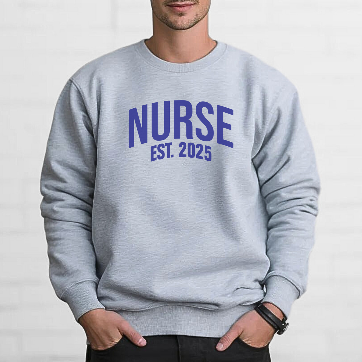 A man wearing a "Nurse Est. 2025 Sweatshirt" in light gray with blue  text, standing against a white brick wall with hands in pockets, looking confident.