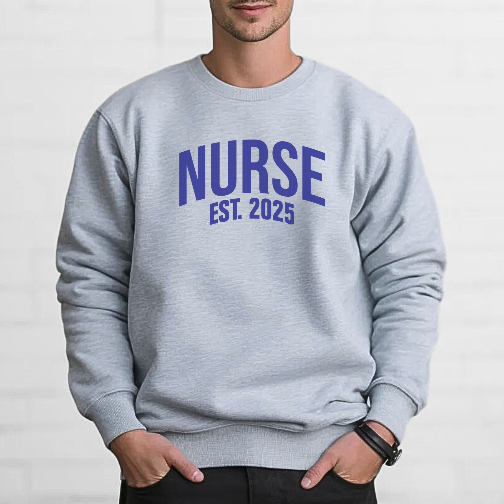 A man wearing a "Nurse Est. 2025 Sweatshirt" in light gray with blue  text, standing against a white brick wall with hands in pockets, looking confident.
