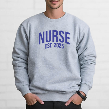 A man wearing a "Nurse Est. 2025 Sweatshirt" in light gray with blue  text, standing against a white brick wall with hands in pockets, looking confident.