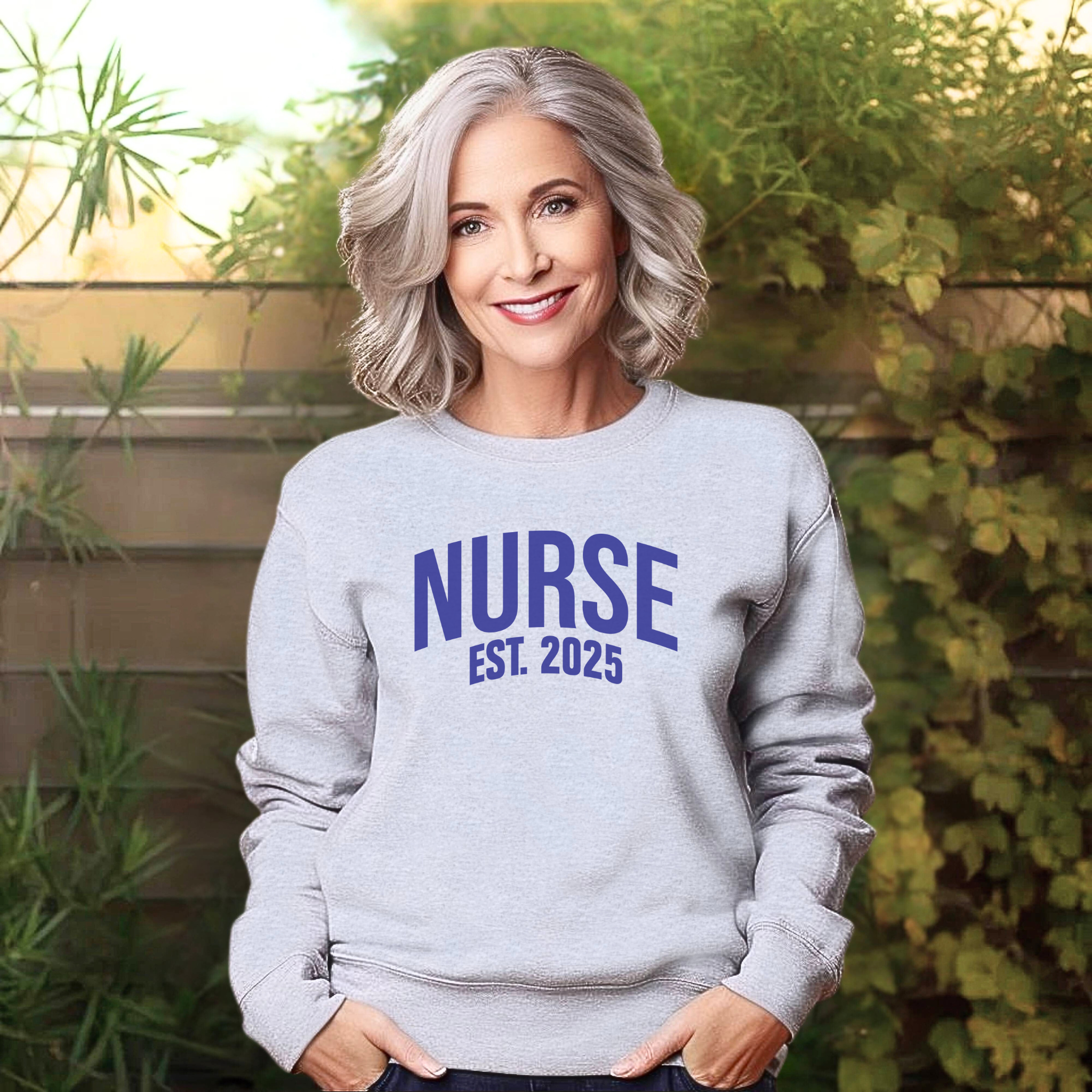 A woman with silver hair wearing a "Nurse Est. 2025 Sweatshirt" in light gray with blue text, standing outdoors with greenery in the background, smiling warmly.
