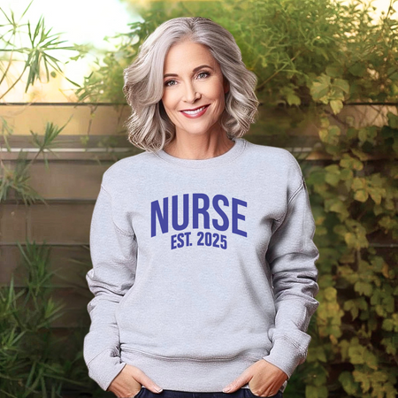 A woman with silver hair wearing a "Nurse Est. 2025 Sweatshirt" in light gray with blue text, standing outdoors with greenery in the background, smiling warmly.