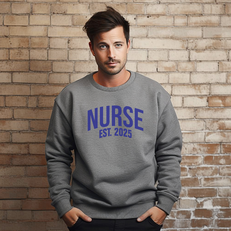 A man wearing a "Nurse Est. 2025 Sweatshirt" in dark gray with blue text, standing confidently against a brick wall, with one hand in his pocket.