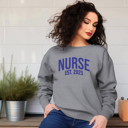 A woman with silver hair wearing a "Nurse Est. 2025 Sweatshirt" in light gray with blue text, standing outdoors with greenery in the background, smiling warmly.