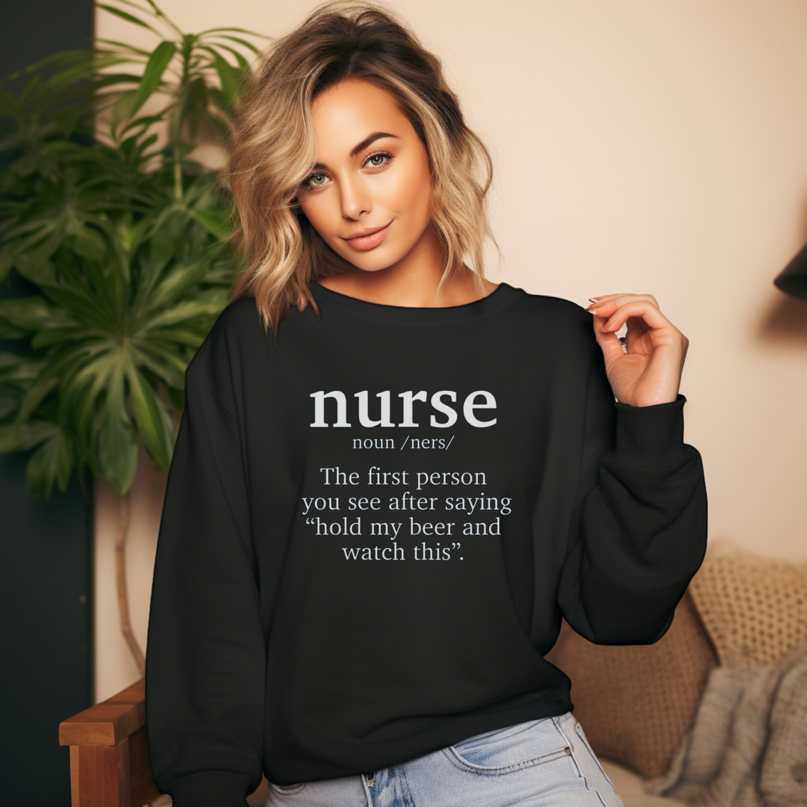 A woman wearing a black sweatshirt with a funny nurse-themed shirt design, reading: 'The first person you see after saying “hold my beer and watch this”.' She is posed indoors with a large green plant in the background.