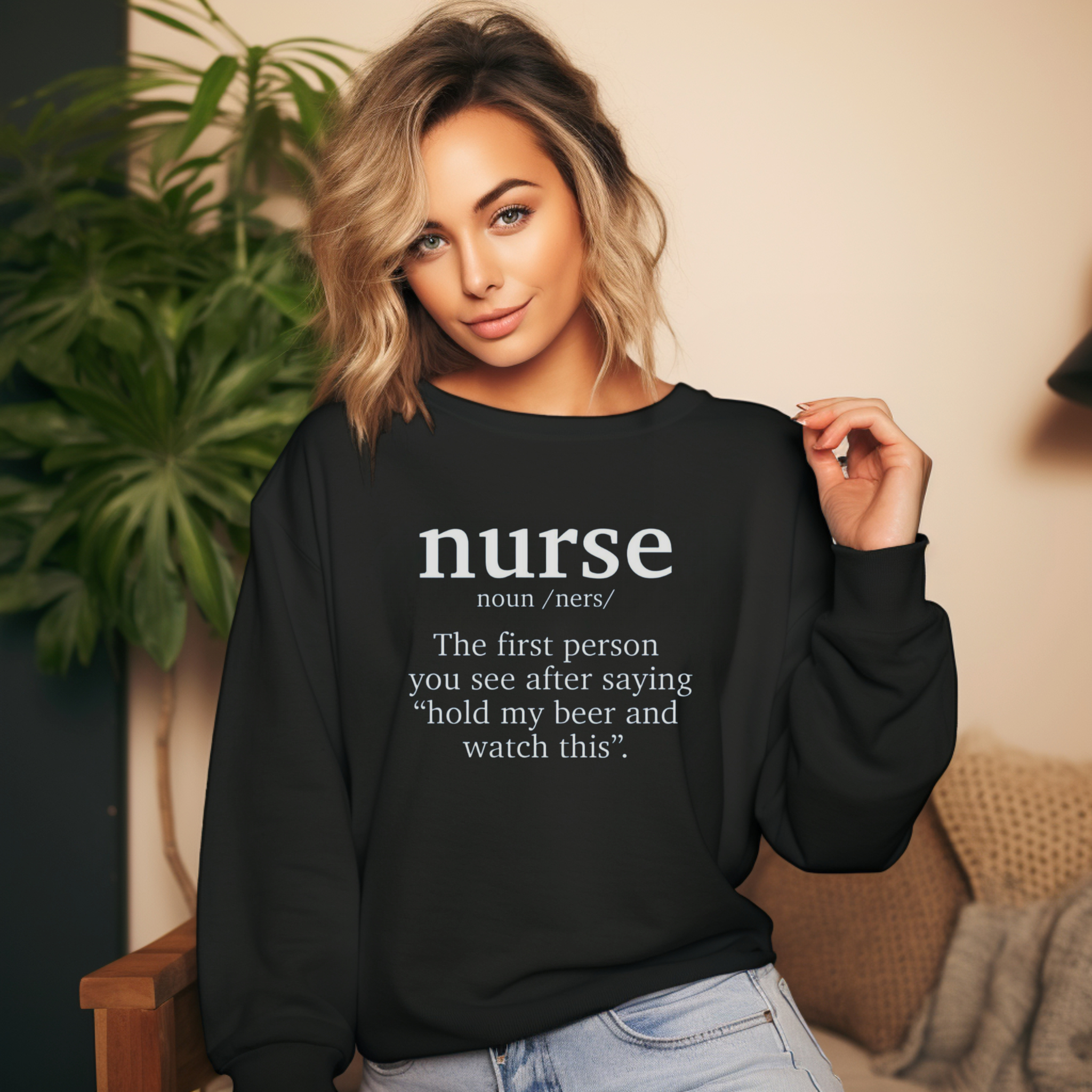 A woman wearing a black sweatshirt with a funny nurse-themed shirt design, reading: 'The first person you see after saying “hold my beer and watch this”.' She is posed indoors with a large green plant in the background.