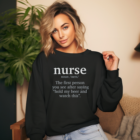 A woman wearing a black sweatshirt with a funny nurse-themed shirt design, reading: 'The first person you see after saying “hold my beer and watch this”.' She is posed indoors with a large green plant in the background.